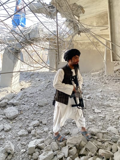 The scene depicts a severely damaged concrete building in Mandisar, Afghanistan, characterized by extensive structural collapse and rubble.
In the foreground, a young male, appearing to be in his late teens or early twenties, stands amidst a dense field of light-colored concrete debris. He wears a black turban, a white long-sleeved tunic, a dark brown plaid vest, and light blue open-toed sandals. He holds a rifle, possibly an M4 variant, in his right hand, positioned vertically with the stock near his hip. His gaze is directed towards the right side of the frame.
In the upper left background, another male figure is partially visible, wearing bright blue loose-fitting pants. He is situated on a higher, extensively damaged portion of the structure, which consists of twisted rebar and fractured concrete slabs.
The environment is dominated by shattered concrete and exposed, bent metal reinforcement bars, indicating significant structural failure. Bright daylight illuminates the scene.