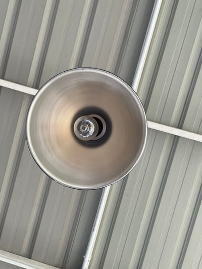 This is a close-up, downward-facing shot of a metallic pendant light fixture with a visible incandescent bulb. The light fixture is suspended from a ceiling made of corrugated metal, painted in a light gray color with alternating raised and recessed bands. Two white pipes or conduits run diagonally across the ceiling, one above and to the left of the light fixture, and another to the right and slightly below it. The image appears to be taken indoors, possibly in a commercial or industrial space, with no indication of the city of Pulokiton, Indonesia, or any people present. The lighting is diffuse, suggesting ambient light rather than direct sunlight.