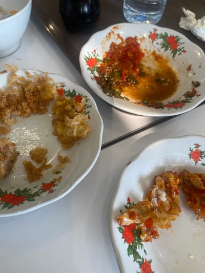 A close-up shot captures the remnants of a meal on a white table, likely in a restaurant setting within Cijantra, Indonesia. Three white, floral-patterned plates are visible, arranged diagonally. The plate in the foreground contains what appears to be crispy fried fish coated in a spicy chili sauce, with some pieces still partially intact. To the left, another plate displays the scattered, fried remnants of what could be vegetables or crispy noodles, along with a small portion of a battered item. The plate further back is almost empty, with only a small amount of chili sauce and chopped chilies remaining. In the background, a white bowl, a dark bottle, and a plastic water bottle are partially visible, suggesting a casual dining environment. The overall scene depicts a recently finished meal, with only leftovers and used dishes remaining.