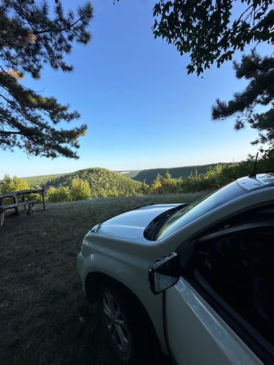 A light-colored SUV is parked on a grassy area with a view of rolling, forested hills under a clear blue sky. The SUV's driver's side door is open, and the side mirror is visible. In the foreground to the left, a weathered wooden picnic table and benches are set up on the grass. Overhead, tree branches frame the scene, casting shadows on the ground. The location is described as being in the city of Pryvorottia Druhe, Ukraine. The image appears to be a static landscape photograph.
