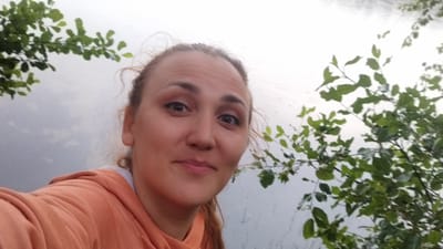 A woman with light brown hair tied back and pulled to the side, wearing an orange hoodie, is captured in a close-up, selfie-style photograph. She is positioned on the left side of the frame, facing the camera with a slight smile. Her eyes are brown, and she has light makeup on. The background is a blurred expanse of greyish water, likely a lake or river, with out-of-focus green foliage from trees or bushes on the left and right sides of the image. The sky appears overcast. The image is not specifically tied to the city of Tarasovskoe, Russia.