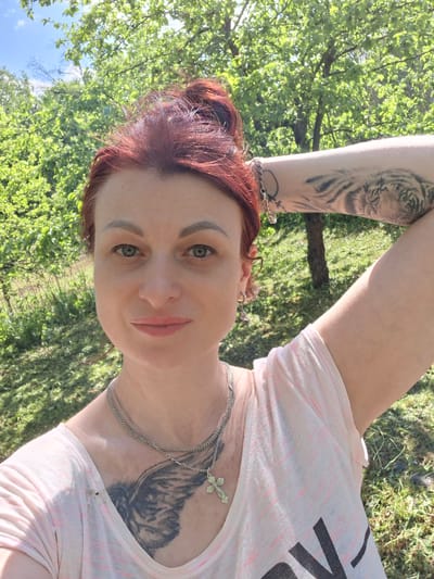 A woman with red hair and green eyes is in an outdoor setting. She has a tattoo on her upper chest, and a tattoo of a tiger on her right arm. She is wearing a light pink shirt and a silver chain with a cross pendant. The background includes green trees and grass, suggesting a park or natural area. The location is Сваткаўскі сельскі Савет, Belarus.