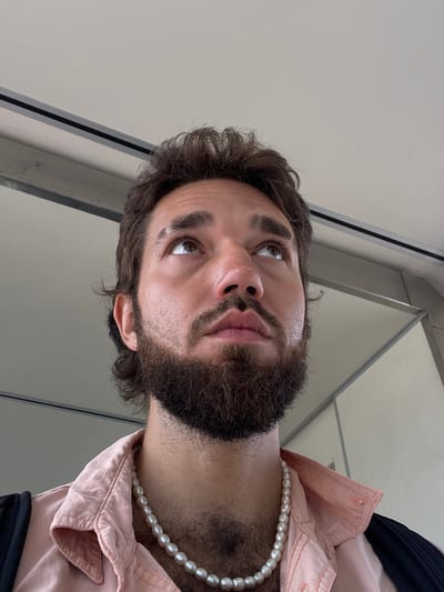 A man with a beard and brown curly hair looks up, his eyes directed towards the top of the frame. He is wearing a light pink collared shirt and a pearl necklace. The background appears to be an indoor environment with a neutral-colored ceiling and walls. The lighting is soft and diffused. The image is a close-up shot, focusing on the man's face and upper torso. There is no visible indication of the man's location in Mai Khao, Thailand, or any other specific city.