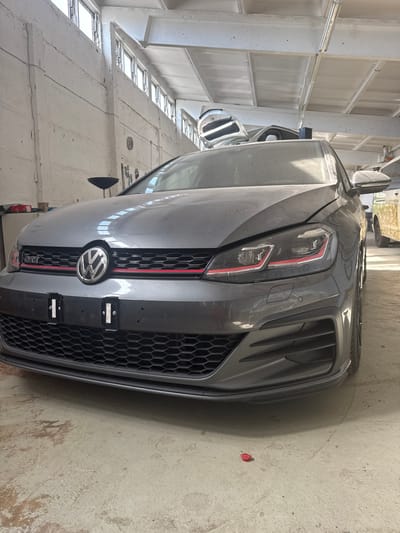 In a dusty garage in Schkopau, Germany, a dark grey Volkswagen Golf GTI is parked. The car, covered in a thin layer of dust, is facing forward, its headlights illuminated with red accents. The car's front grille, with the iconic Volkswagen emblem and "GTI" badge, is clearly visible. Behind the car, parts of what appear to be other vehicles or car parts are stacked. The garage has a concrete floor and walls, with a high ceiling featuring exposed beams and industrial lighting. The overall impression is one of a workshop or storage area.