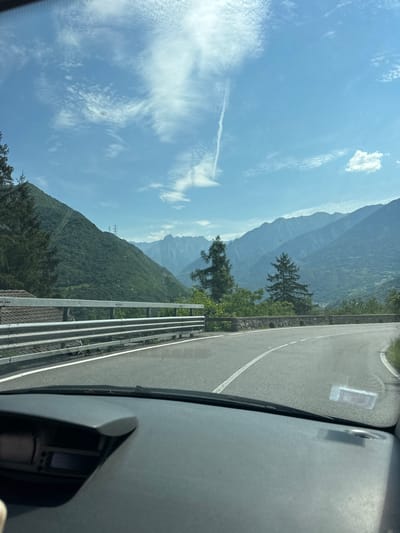 The image captures a scenic view from a vehicle driving on a winding road in Malegno, Italy. The road curves to the right, bordered by a metal guardrail on the left. Beyond the guardrail, a residential building with a tiled roof is visible. The landscape is dominated by lush green mountains, with tall evergreen trees dotting the slopes. In the distance, majestic, hazy blue mountains form the backdrop under a clear blue sky with scattered white clouds, including a distinct contrail. The foreground shows the dashboard of the vehicle, with a partial view of the instrument panel. Power lines and transmission towers are also visible on the mountainside. The overall impression is one of a serene, mountainous region on a sunny day.