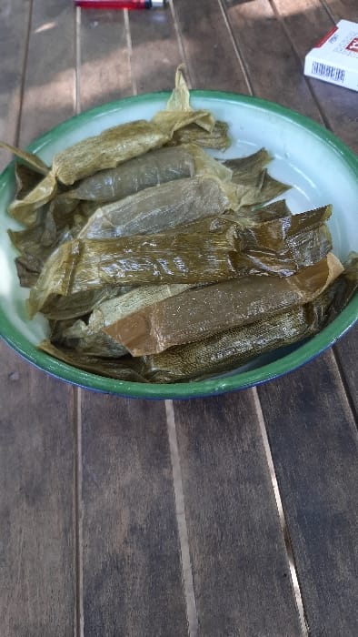 A light green enameled metal bowl with a darker green rim contains approximately 15-20 elongated food items. Each item is individually wrapped in what appears to be olive-green to brownish natural leaf material, exhibiting a somewhat translucent and wrinkled texture. The bowl is positioned on a dark brown wooden plank table with visible grain and seams. In the upper left quadrant of the frame, a red disposable lighter is partially visible on the table. In the upper right quadrant, a white cigarette pack with red accents is also partially visible on the table. Direct sunlight illuminates the scene, evidenced by distinct shadows on the wooden surface. The location is RawangItiek, Indonesia.