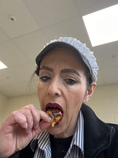 A woman with dark hair and heavy makeup is holding a piece of food, covered in a dark sauce and colorful sprinkles, close to her mouth. She is wearing a white cap with a lace trim, a black shirt and jacket, and a white and black striped shirt underneath. The background is a white tiled ceiling with fluorescent lighting. The location cannot be determined as Mensdorf, Luxembourg.
