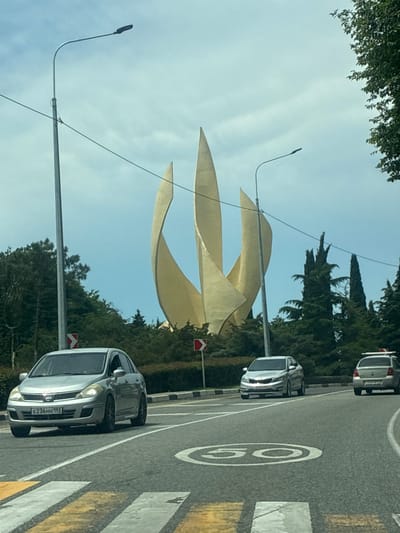 This is a daytime, eye-level, exterior shot of a road in Novomikhaylovsky, Russia, with a large golden monument in the background. The monument is abstract in design, resembling three tall, curved, petal-like structures converging at the top. It is surrounded by lush green trees and foliage. A tall lamppost stands to the left of the monument, with utility wires stretching across the frame.

The road in the foreground has clear lane markings and a crosswalk. Several cars are visible: a silver sedan in the left lane facing forward, a silver hatchback in the right lane also facing forward, and a white car with a roof rack further down the right lane. A speed limit sign indicating "50" is painted on the road surface. The sky is overcast with visible clouds. Two red and white directional traffic signs are also present on the side of the road. The overall scene depicts a public road with a prominent landmark.