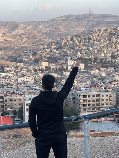 The image displays a panoramic view of the city of Qudsaiya, Syria, seen from an elevated perspective. In the foreground, a lone individual stands with their back to the viewer, facing the sprawling urban landscape. The person is wearing a black hooded long-sleeved top and black trousers. Their right arm is extended straight upwards and slightly to the right, with the hand open. A dark, weathered metal railing runs horizontally across the lower part of the frame, separating the rocky, unpaved foreground from the cityscape.

The midground is dominated by a densely packed residential area built into hillsides. Numerous multi-story buildings, predominantly light in color (beige, grey, white), feature visible rooftops, many equipped with red or blue water tanks. Interspersed among the buildings are sparse patches of green vegetation. To the lower right, a large, light-colored paved area resembling a basketball court is visible, with several small, dark figures appearing to be playing on it. In the left-midground, two tall, slender industrial chimneys stand against the distant hills.

The background consists of arid, rolling mountain ranges, receding into a light haze. On a prominent peak in the far right background, a cluster of communication towers is discernible. The sky above is a pale blue with some subtle cloud formations. The lighting suggests daytime conditions, possibly late afternoon.
