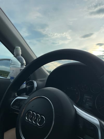 The image shows a close-up view from inside a car, looking out towards a cloudy sky with the sun partially visible. The dominant object is the steering wheel, which has the Audi logo embossed on it. A digital display on the dashboard is partially visible, showing "00200928" and "300 km". A partially full plastic water bottle with a green cap is placed on the steering wheel. The setting appears to be daytime, with bright but overcast weather. The orientation of the image is such that the steering wheel is on the right side of the frame, suggesting the viewpoint is from the passenger seat or the car is in a country where driving is on the right. The location is identified as Vadul lui Voda, Moldova. No people are visible, and the overall mood is neutral, focusing on the car interior and the external environment.