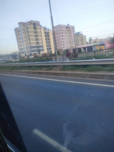 The image depicts an urban landscape in Theta ward, Kenya. In the foreground, a dark asphalt road surface with a single white lane marker is visible, suggesting it was captured from a moving vehicle. A concrete curb separates the road from a grassy median strip, which is protected by a silver metal crash barrier.

Beyond the barrier, a pedestrian area or shoulder runs parallel to the road. Several indistinct human figures are present, with one individual in red attire walking towards the right. A tall, slender utility pole extends vertically from this midground section. Behind the pedestrian area, a dark fence separates it from a series of single-story commercial buildings, some featuring yellow and orange-red facades. A white vehicle and a large dark vehicle (possibly a bus) are partially visible further back on the left.

The background is dominated by multiple multi-story residential buildings. On the left, a building identified by "WALES PLACE" signage displays a facade with alternating vertical sections of beige, light blue, and grey. To its right, a prominent pink-colored building, featuring balconies and structural elements, is marked with "JALUO HEIGHTS" and "JALUO RESIDENTS" at its apex. Both primary buildings appear to be approximately 10-12 stories high. Additional, less distinct residential structures and sparse vegetation are visible in the distant background. Overhead, multiple utility lines or power cables are stretched across the sky, which is a light blue, hazy tone indicative of daytime conditions.