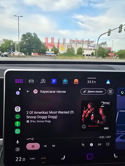 The image shows a car's infotainment system display, likely in Bischofsheim, Germany, as indicated by some German text on the screen and the external scene. The main subject on the screen is a music player displaying album art for "2 Of Amerikaz Most Wanted" by 2Pac ft. Snoop Dogg. The song is currently playing, with the progress bar showing 0:20 out of 4:07. Other music player controls like play/pause, skip forward, and repeat are visible.

The screen also shows various icons and information:
- A temperature reading of 33.5°C.
- A "MODE" button, an "ASSIST" button, and a "P" (parking) button.
- A "Джем сешън" (Jam session) icon.
- A toggle for "A OFF" (likely automatic engine start/stop).
- A row of app icons at the bottom, including Google, Spotify, Phone, and WhatsApp.
- "CLIMA" and "AUTO" buttons, suggesting climate control features.

The external scene visible through the car window features a bright, cloudy sky, suggesting daytime. There are buildings with signage that includes "Me Via Markt" and "Fink". There are also traffic lights and streetlights, indicating a road or a commercial area. The overall impression is a modern car interior with a functional and information-rich display, with the user potentially driving or parked in a commercial area.