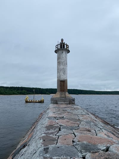 A lighthouse stands tall on a stone pier, overlooking a body of water. The lighthouse is cylindrical and weathered, with a simple wooden door at its base and a lantern room at the top. A boat with a dragon-shaped prow and stern, adorned with gold, floats in the water to the left of the pier. The background features a dense forest of green trees lining a sandy shore, under a cloudy sky. The pier itself is constructed from large, rough-hewn stones in varying shades of gray and pink. The scene appears to be set in a natural, somewhat remote location, likely Моторное, Russia, given the presence of the lighthouse, water, and forested shoreline.