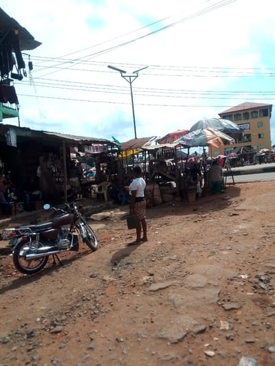 The image shows an open market scene in Orlu, Nigeria. The ground is a dusty, reddish-brown dirt, cracked and uneven. A woman stands in the foreground, wearing a white shirt and patterned skirt. A brown and silver motorcycle is parked nearby. Several makeshift stalls with corrugated iron or tarpaulin roofs are clustered together, selling goods. People are seated and standing around the stalls, engaging in commerce. A street light pole rises from the center of the market area. In the background, a multi-story building with orange roofing is visible on the other side of a street with cars on it. Overhead, electrical wires crisscross against a sky with scattered clouds.