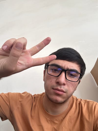 A young man with short black hair, wearing black-rimmed glasses and an orange t-shirt, is looking directly at the camera. He is holding up his left hand in a peace sign, with his fingers extended. His skin has some redness and blemishes. The background is a plain, light-colored wall. There is no indication of the city of Danghara, Uzbekistan, in this image.