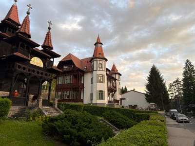 The image shows the exterior of two buildings in Sovata, Romania. The building on the left is a dark wooden structure with red-tiled turrets topped with crosses, and a mosaic under the roofline. A stone staircase leads up to its entrance. Adjacent to it, to the right, is a multi-story building with white and brown exterior walls, also featuring a red-tiled roof and turrets. The buildings are surrounded by green bushes and grass. Further down the street to the right, several parked cars are visible, and a white building can be seen in the background along with tall evergreen trees. The sky is overcast with light visible between the clouds. The photograph is taken from ground level.