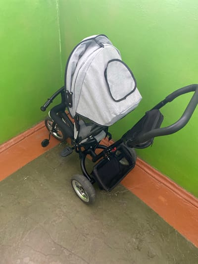 An indoor, high-angle shot shows a gray and black baby stroller parked in a corner where two walls meet. The wall on the left is painted a bright green, and the wall on the right is painted a bright orange at the bottom and green at the top. The floor is a mottled green and brown. The stroller is a three-wheeled model with a gray fabric canopy and a black fabric storage bag. A black handle is visible at the top of the stroller. The stroller appears to be empty and stationary. The scene suggests a domestic or communal indoor space within a building in the Beskol rural district of Kazakhstan.