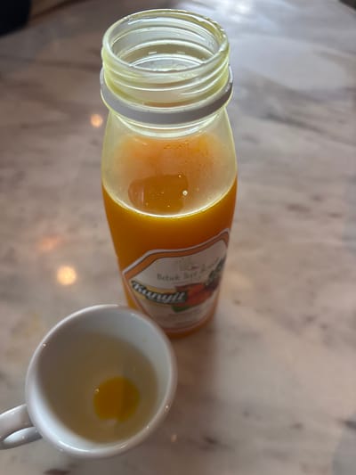 A glass bottle of Kunyil brand "Bebek Tepi Sawah" mango juice lies on its side, mostly full. A small white mug sits next to the bottle, containing a small amount of the same orange juice. The bottle's white cap is open, revealing the bottle's opening. The background is a marble surface with blurry light spots. The focus is on the bottle and mug, highlighting the juice.