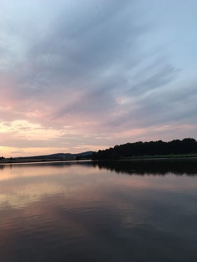 The image depicts a serene lake at dusk, with the sky painted in soft hues of pink and blue. A line of dark trees forms the horizon on the far side of the lake, silhouetted against the fading light. The water's surface is calm, reflecting the sky and the distant treeline, creating a mirrored effect. The location is identified as the Kestřany village, Czechia. The scene is devoid of any people or discernible activity, emphasizing the tranquility of the natural landscape.