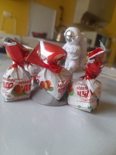A close-up, eye-level shot captures three wrapped candies and a white astronaut figurine on a white, textured surface. The candies are individually wrapped in shiny red and white foil with images of hazelnuts and text in a foreign language, suggesting they are a confectionery product. The astronaut figurine, located behind the candies, is white with a reflective silver helmet. The background is blurred, showing hints of a kitchen environment with yellow walls and appliances. The overall scene appears to be indoors, likely in a domestic setting. There is no discernible indication of the city of Salsk, Russia, or any specific location beyond a general indoor setting.