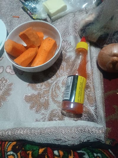 The image captures a close-up, top-down view of several food items and kitchen essentials arranged on a patterned tablecloth, likely on a dining or kitchen table.

In the center-left, a white, round bowl holds several pieces of peeled, thick-cut orange carrots. There are at least five pieces visible, suggesting they are prepared for cooking or eating.

To the right of the bowl, a small, clear plastic squeeze bottle containing an orange-red sauce stands upright. The bottle has a yellow plastic cap with a red tip. A yellow label wraps around the bottle, displaying a black barcode and text primarily in Cyrillic script. Although difficult to read perfectly due to resolution and angle, parts of the text are visible, including what appears to be ingredients or product description such as "СОСТАВ: ПРИПРАВА, ПЕРЕЦ, КРАСНЫЙ, ПИЩЕВАЯ, СЛАДКИЙ" (Composition: seasoning, pepper, red, food, sweet).

Further to the right, a whole, unpeeled brown onion is partially visible. In the upper background, slightly to the left of the onion, a rectangular piece of light yellow or white cheese is visible, enclosed in clear plastic wrapping. Another small, orange sliver, possibly a piece of carrot or carrot peel, is seen near the cheese. A corner of a white plate is visible on the far left.

The items rest on a light beige or off-white tablecloth featuring an ornate, repeating pattern in a muted gold or bronze color. Below the tablecloth, in the bottom portion of the image, a colorful, intricately patterned rug or textile with shades of red, green, orange, and black is visible.

The lighting appears to be indoors and somewhat dim, casting subtle shadows from the bowl and bottle. The overall scene suggests a moment during food preparation or a casual meal setting.