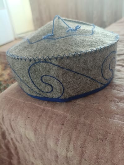 A close-up, eye-level, slightly high-angle shot shows a gray felt hat with blue decorative stitching and a blue trim. The hat is cylindrical with a rounded top and is placed on a pink quilted surface. The hat has a decorative blue embroidered swirl pattern on its side and blue stitching along the top edge. There are also loose blue threads extending from the top of the hat. The background is blurred, showing what appears to be a white vertical object and a glimpse of a patterned rug. The image is likely taken indoors, possibly in a residential setting in Zhanaozen, Kazakhstan, given the context provided. No people are visible in the image, and no specific actions or interactions are depicted. The focus is solely on the hat and its immediate surroundings.