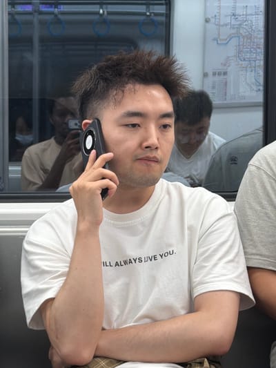 A man is seated on a subway, holding a smartphone to his ear, and looking forward. He has short, spiky dark hair, and is wearing a white t-shirt with black text that reads "ILL ALWAYS LOVE YOU.". He is wearing plaid shorts. Behind him, two other individuals are visible through the subway window, also appearing to be passengers. A map of the subway system is displayed on the wall behind them. The environment suggests a public transportation setting.