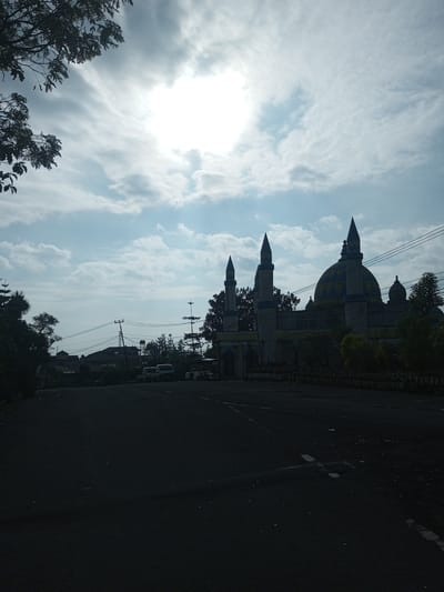 The image shows the Masjid mosque in Mya Sit Kaing Ywar Thit, Myanmar. The mosque has a central dome and three minarets. It is silhouetted against a cloudy sky with a bright sun obscured by the clouds. In the foreground, there is a dark asphalt road. Across the road, there are several vehicles, a few buildings and utility poles are visible in the background. Trees and vegetation flank both sides of the road and mosque.