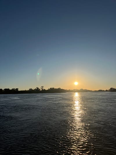 The image shows a sunset over the Zambezi River in Victoria Falls, Zimbabwe. The sun is positioned in the center of the frame, casting a bright glare across the water. The sky is a clear blue, fading to a pale yellow near the horizon. On both sides of the river, there are trees on the riverbank. The water is dark, with ripples and waves reflecting the sunlight.