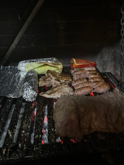 The image displays a close-up, elevated view of a variety of food items cooking on a charcoal grill within an enclosed, dark environment, indicative of a traditional parrilla in Villa San Andrés, Argentina. The grill grate, visibly marked by use and ash, is positioned above glowing red embers. On the left, two items are wrapped in aluminum foil; the upper item features exposed green corn husks, suggesting a corn-based preparation. To the right of these, two pieces of light-colored meat, consistent with chicken or thin pork cuts, are browning. Adjacent to these, two parallel rows contain approximately twelve sausages, exhibiting a cooked, reddish-brown exterior. A large, thick cut of red meat, possibly beef, occupies the lower right section of the grill. The background shows dark, brick-like walls, with a diagonal metal beam in the upper left and a metal chain hanging vertically on the far right, likely part of the grill's height adjustment mechanism. No individuals are present.