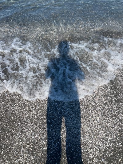 A high-angle shot captures a shadow of a person standing on a beach of small, light-colored pebbles and dark sand. The shadow, cast by the sun from the left, stretches towards the viewer, obscuring the ground. In the upper portion of the frame, the shallow, clear water of the sea laps onto the shore, creating a line of white foam and bubbles that partially covers the pebbles. The water surface reflects sunlight, creating a shimmering effect. The scene is likely from Shekvetili, Georgia, known for its pebble beaches.