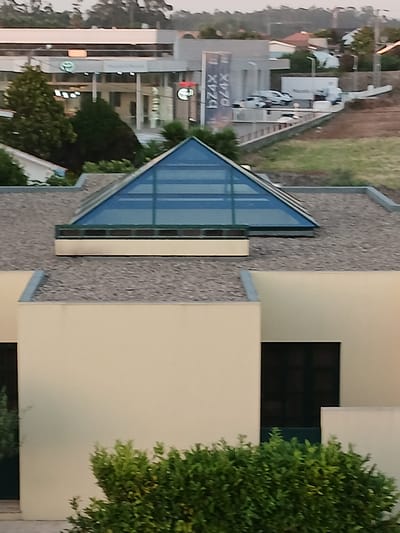 The image captures a section of a building with a distinctive pyramidal glass skylight situated on a gravel-covered roof. The building's facade is painted a pale yellow, and dark rectangular windows are visible. In the background, a Toyota dealership with prominent signage featuring the Toyota logo is discernible, along with several vehicles parked outside. The scene is set against a backdrop of trees and a hilly landscape, suggesting a suburban or semi-rural environment in Póvoa de Varzim, Portugal. The lighting indicates it is likely late afternoon or early evening.