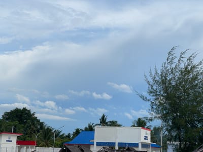The image captures a bright, cloudy day in Lancok, Indonesia. In the foreground, a white building with red trim and the word "BENGKEL" in blue lettering is visible. Adjacent to it is another building with a striking blue roof. Beyond these structures, lush green trees and palm trees dominate the landscape. On the right side of the frame, a large, dense evergreen tree with thin, feathery branches stands prominently against the sky. The sky itself is filled with a mix of white and grey clouds, suggesting a dynamic weather pattern. The scene is devoid of people, presenting a still and serene environment.