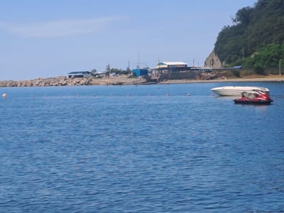 The image depicts a coastal scene in Dzhubga, Russia, likely a resort area. The foreground is dominated by calm blue water with gentle ripples, suggesting a settled day. Several small orange buoys are visible, marking a boundary or safety zone in the water. In the mid-ground, two watercraft are moored: a white motorboat with dark trim, and a red and black jet ski. These are situated in the sea, close to shore.
The background reveals a rocky shoreline topped with a variety of structures. These appear to be small buildings, possibly part of a beachfront settlement or tourist facilities, with roofs of various colors, including green and yellow. Some of these buildings are positioned on a raised embankment constructed from large stones. Further back, a steep, tree-covered hill rises, characteristic of the Black Sea coast. The sky is a clear, pale blue with no clouds. The overall impression is a tranquil summer day at a seaside location.