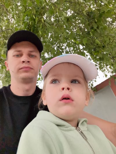 In an outdoor, low-angle shot, a man and a young child are positioned in the foreground. The man, on the left, wears a black baseball cap and a black t-shirt, looking directly at the camera with a neutral expression. The child, on the right, is closer to the camera, wearing a pastel-colored baseball cap and a light green hooded jacket. The child's gaze is directed upward and to the left, with an open-mouthed expression. The background consists of lush green foliage from trees overhead, with a sliver of a building visible on the right. The lighting is diffused, suggesting an overcast day or shade. The scene is a close-up portrait, with the subjects dominating the frame.