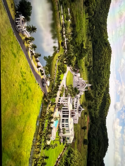 An aerial view captures a large, sprawling complex of white buildings with domes, situated amidst lush green landscaping and palm trees. A body of water, possibly a lake or reservoir, is visible to the left, reflecting the sky. A paved road winds through the property, leading to various structures. The complex appears to be a resort or a place of worship, characterized by its symmetrical architecture and manicured grounds. In the background, a densely forested hillside rises, under a sky with scattered clouds. The image is vertically oriented and appears to be captured from a drone or helicopter. The location is identified as Vientiane Capital, Laos.