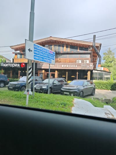 The image shows a street view of a building in Glukhovo, Russia. The building appears to be a restaurant or a multi-purpose establishment with a wooden facade and large windows. Several cars are parked in front of the building. A street signpost with directional arrows and text in Russian is visible, indicating destinations like "ИЛЬИНСКАЯ БОЛЬНИЦА" (Ilyinskaya Hospital), "НИКОЛЬСКАЯ СЛОБОДА" (Nikolskaya Sloboda), and "НИКОЛО-УРЮПИНО" (Nikolo-Yuryupino). The building itself has signage that reads "Якитория" (Yakitoria) on the left and "МИШПАХА" (Mishpaha) and "Village KITCHEN" on the right. The sky is overcast, suggesting cloudy weather. The overall scene depicts a commercial area on what seems to be a typical day. No people are clearly visible in the image, with the focus being on the architecture, vehicles, and signage.