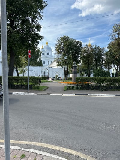 A sunlit, exterior medium shot captures a street scene in front of a white building with gold-domed roofs, likely a church or monastery, in Putyatino (Semivragovskaya S/A), Russia. The building is partially obscured by lush green trees. In the foreground, a gray asphalt road runs horizontally across the bottom of the frame. To the left, a portion of a silver car is visible. A gray metal pole stands prominently on the left side of the image.
A red and white "No Entry" sign is mounted on a pole to the left of a manicured green hedge. Behind the hedge, a person is walking towards a gated entrance of the white building. Several cars are parked or driving near the entrance. A bust monument on a pedestal is situated in front of the building, surrounded by a flower bed with bright orange flowers. The sky is blue with scattered white clouds.