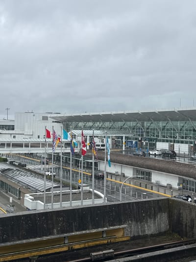 The image shows a view from an elevated train or monorail looking down at an airport or a large transportation hub. The scene is outdoors, with a cloudy and overcast sky, suggesting a cool or rainy day. The architecture of the buildings appears modern, with large glass facades and metallic structural elements.

In the foreground, we see the railing of the elevated track, with some dirt and debris on it. Below the track, there are several flags flying from poles. These flags appear to represent different countries or regions, with some recognizable patterns like the Canadian flag (red and white maple leaf) and a flag with red and white stripes and a blue canton (possibly the US flag). There are also some blue and white flags, and one with a red and yellow pattern.

Beyond the flags, there is a long, modern building with a glass-fronted section and a white facade. There are signs and windows visible on this building. Cars are visible parked or driving along a road or drop-off area adjacent to the building. Some people are visible, but they are small and indistinct due to the distance.

To the right of the building and track, there is a dark, textured surface, possibly a concrete wall or embankment, with some green vegetation growing in parts. Further to the right, there are train tracks or some kind of guideway with yellow markings.

The overall impression is of a busy, functional transportation infrastructure on a dull day. The presence of multiple national flags might indicate an international airport or a location that celebrates diversity. There is no discernible text that is clear enough to read. The scene is static, with no specific event clearly depicted, but it captures the atmosphere of a transit point. The location is identified as Richmond, Canada.