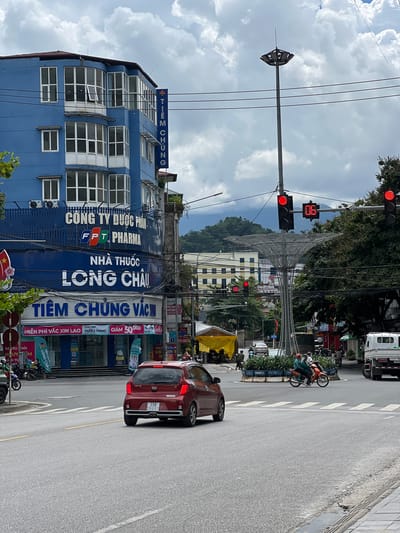 This is a street-level, eye-level shot of an intersection in Hà Giang 2 Ward, Vietnam. The scene is dominated by a large, bright blue building on the left, which has multiple floors and many windows. A vertical blue sign with white text reading "TIEM CHUNG" is affixed to the building's facade. Below this, larger white text reads "NHA THUOC LONG CHAU." Further down, another blue sign with white text reads "TIEM CHUNG VAC."
On the right side of the image, a tall street light stands prominently, supporting traffic signals and a digital display showing "06." There are also trees and other buildings visible in the background. The street is paved with asphalt, and there are crosswalk markings.
In the foreground, a red Kia car is driving away from the viewer, its license plate reading "23A 059.51." Several motorcycles are also present, some parked and others in motion. People are visible on the motorcycles and walking on the sidewalk. A white truck is parked on the far right. The sky is partly cloudy. The overall atmosphere suggests a busy urban environment.