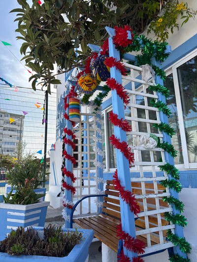The image depicts an outdoor scene in Costa da Caparica, Portugal, featuring a blue-painted wooden trellis structure adorned with festive red, green, and white tinsel garlands. A wooden bench is integrated into the trellis. Above and to the left, green foliage from a tree with large leaves is visible. Several colorful, paper pinwheel-like decorations and a cylindrical, multi-colored object are also suspended from the structure. The trellis is situated next to a white building with blue trim and a window. In the foreground, two blue planters with white stripes contain plants. A wire fence is visible behind the planters, with small, triangular flags strung along it. In the background, a tall, light-colored building stands against a cloudy sky. A mural depicting several figures is partially visible on the white wall to the left of the trellis.