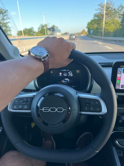A person is driving a Fiat 500 in Mola di Bari, Italy. The driver's hand is visible on the steering wheel, sporting a wrist watch with a brown leather strap. The digital dashboard displays a speed of 76 km/h and the car's odometer reads 1994 km. A smartphone is mounted on the dashboard showing a navigation app. The driver's leg is also visible. The outside view through the windshield shows trees, other vehicles, and a bright, sunny sky, suggesting daytime. The temperature is 27°C. The steering wheel has controls for phone, audio, and cruise control.