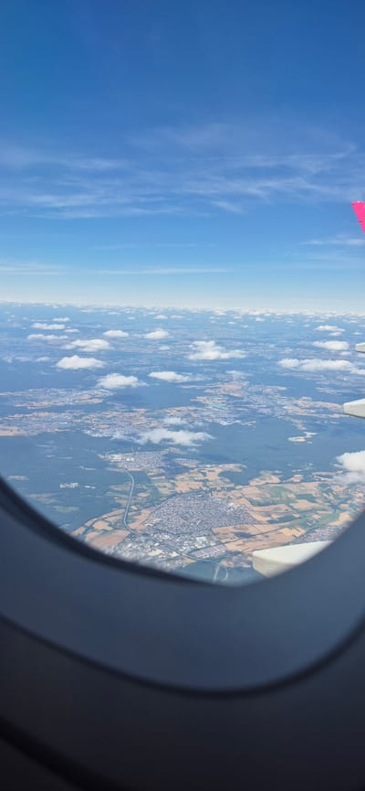 An aerial view from an airplane window shows a landscape of scattered clouds over a vast expanse of land. Below the clouds, a mosaic of agricultural fields, forests, and urban areas stretches to the horizon. A highway is visible, cutting through the landscape and connecting various settlements. The airplane's wing, painted pink at the tip, is visible in the upper right corner of the image, indicating the perspective is from inside the aircraft. The sky is a clear blue with streaks of white clouds. The scene appears to be over the city of Spata-Loutsa Municipal Unit, Greece, given the characteristic urban and agricultural layout.