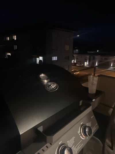 The image depicts a nighttime scene from an elevated vantage point in Höri, Switzerland, likely a balcony or upper floor residence. In the immediate foreground, the upper portion of a black gas grill is visible, featuring a circular temperature gauge and an oval metallic emblem engraved with "Barbecue Grill Club" on its closed lid. The lower right portion of the grill shows part of its control panel with at least two prominent metallic control knobs. To the right of the grill, resting on a dark surface, is a clear plastic cup with a lid and straw, containing a light-colored liquid and ice.
The midground reveals a residential environment under artificial illumination. A multi-story building stands in the left-center, presenting a dark facade with several illuminated windows on different floors; one lower window prominently displays a dark, indistinct silhouette, potentially a human figure. Further back and to the right, another multi-story building is visible, illuminated by external facade lighting and internal window lights. A paved area, consistent with a street or parking lot, is discernible between the buildings, where at least one light-colored vehicle is parked. The scene is illuminated by multiple artificial light sources, including streetlights or building-mounted lights, which cast bright reflections and deep shadows across the structures and ground. The background consists of a dark night sky. No explicit actions or interactions are clearly discernible beyond the overall environmental setting.