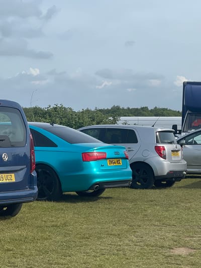 The image captures a row of parked cars on a grassy field, likely in the city of Sevenoaks, United Kingdom. The cars include a blue Vauxhall, a teal Audi A3 with license plate OV64 HSE, a silver hatchback, and a white van. The background consists of a treeline against a partly cloudy sky. The scene appears to be outdoors during daylight hours.