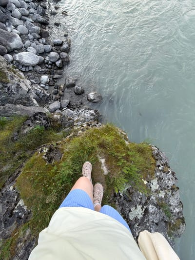 The image depicts a first-person perspective looking down at a person's legs and feet standing on a rocky, moss-covered outcrop next to a body of water. The person is wearing blue and white striped shorts and athletic shoes. The water appears to be a river with a light blue-green hue and visible ripples. The background consists of a rocky shoreline with large grey boulders and a grassy embankment. The location is not identified as Uznezya, Russia; no specific geographical or urban features are visible to confirm this.
