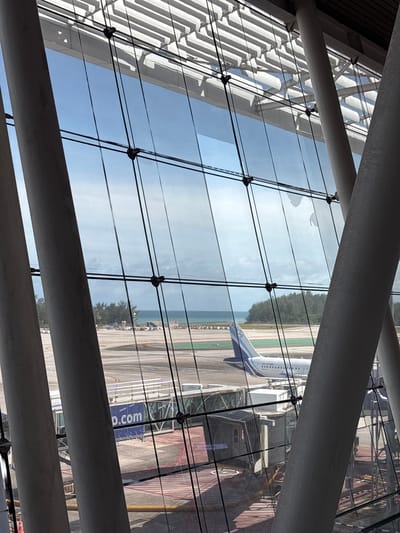 The image depicts a panoramic view from within an airport terminal, looking outwards onto the tarmac, an aircraft, and a distant coastal landscape. The scene is illuminated by natural daylight.
In the immediate foreground, large, inclined, cylindrical grey columns of the terminal's structure frame the view. A grid-like system of black vertical and horizontal tension cables or rods, secured by visible connectors, supports the expansive glass facade. Reflections of the terminal's white, slatted ceiling structure are visible in the upper portion of the glass.
The midground features an airport tarmac. An IndiGo airline aircraft, identifiable by its blue and white tail with the airline's logo, is partially visible. The fuselage is white with a row of windows and the partial registration "VT-". A grey and blue jet bridge is extended towards the aircraft, displaying the partial text "p.com" in white letters on its side. Various ground markings are visible on the tarmac.
In the background, beyond the tarmac, a dense line of green trees separates the airport grounds from a sandy coastline. Further in the distance, a body of water, likely the Andaman Sea, extends to the horizon under a light blue sky with scattered white clouds. The location is identified as ตำบล ไม้ขาว (Mai Khao), Thailand, consistent with the coastal airport environment, specifically Phuket International Airport (HKT).
