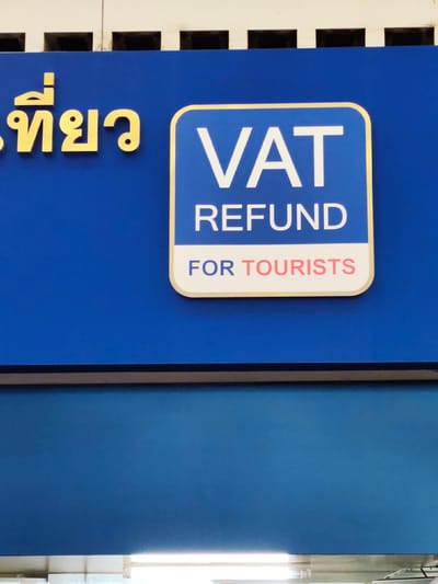 The image shows a close-up of a blue sign in Tambon Mai Khao, Thailand. The sign has yellow Thai lettering on the left, which appears to say "เที่ยว" (Tîao), meaning "travel" or "tour". To the right of the Thai text is a rounded rectangle with a yellow border, containing white English text. The top line inside the rectangle reads "VAT", and the second line reads "REFUND". Below that, in pink text, it says "FOR TOURISTS". The sign is mounted on a blue background. Above the sign, there is a white structure with dark rectangular openings. Below the sign is a darker blue surface.