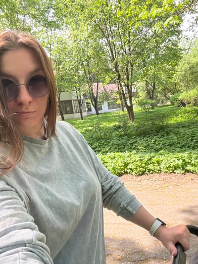 A selfie shows a woman with brown hair and sunglasses. She is wearing a light green sweatshirt and holding a stroller. In the background are trees and a building. The location is assumed to be Tokkari-Lend, Russia.