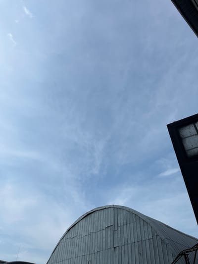 The image, taken from a low-angle perspective, shows a sky filled with light, wispy clouds against a pale blue background. Two dark, angular structures frame the sky. On the left, a portion of a dark, flat roof or overhang is visible. On the right, a large, curved section of a corrugated metal structure dominates the view, its metallic surface reflecting the ambient light. The scene suggests an outdoor setting, possibly urban or industrial, given the architectural elements. The lighting and cloudy sky indicate it is daytime, likely overcast or with scattered clouds. There is no visible text in the image. The absence of people and any indication of specific activity allows for a focus on the architectural forms and the atmospheric conditions. The overall mood is neutral and contemplative, emphasizing the interplay of man-made structures and the natural sky.